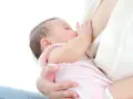 Malutkie dziecko karmione piersią. Mama delikatnie przytrzymuje główkę, a dziecko chwyta pierś. Nie widać kapturków na brodawki.