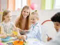 Uśmiechnięta opiekunka pomaga dzieciom malować. Warto wiedzieć, ile zarabia opiekun w żłobku, bo to praca pełna pasji.