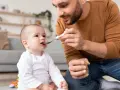 Tata karmi dziecko z słoiczka, używając łyżeczki do rozszerzania diety. Maluch z zaciekawieniem patrzy na jedzenie.