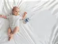 Uroczy noworodek leży na białej pościeli obok grzechotki. Rodzice zastanawiają się, jakie pieluszki dla noworodka: tetrowe czy bambusowe?