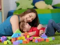 Uśmiechnięta opiekunka bawi się z dzieckiem wśród kolorowych klocków. Zastanawiasz się, ile kosztuje opieka nad dzieckiem za godzinę?