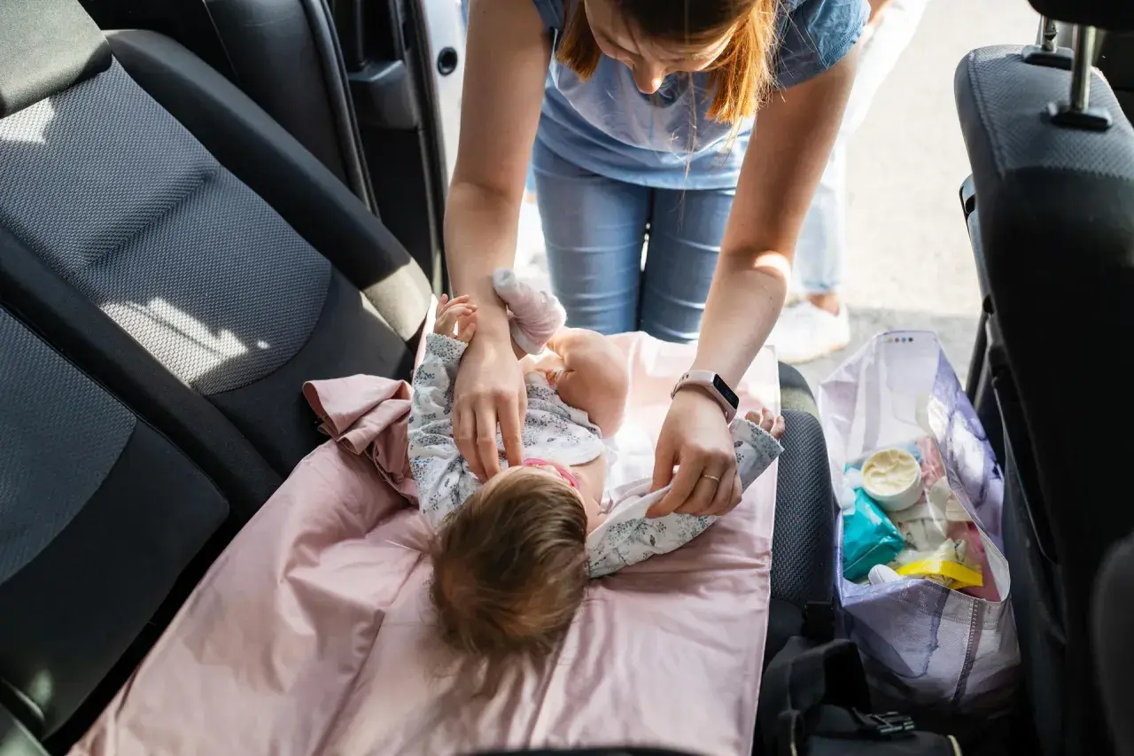 Mama uczy się, jak przebierać noworodka w samochodzie. Na różowej macie leży dziecko, a obok torba z akcesoriami.