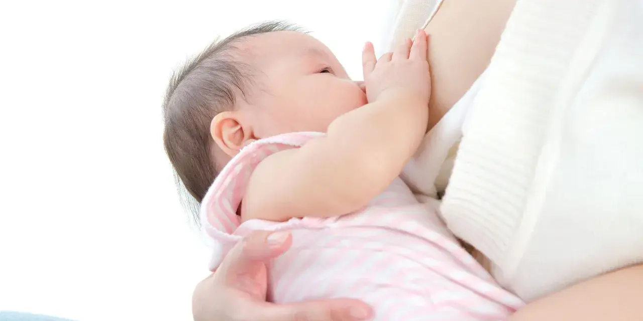 Malutkie dziecko karmione piersią. Mama delikatnie przytrzymuje główkę, a dziecko chwyta pierś. Nie widać kapturków na brodawki.
