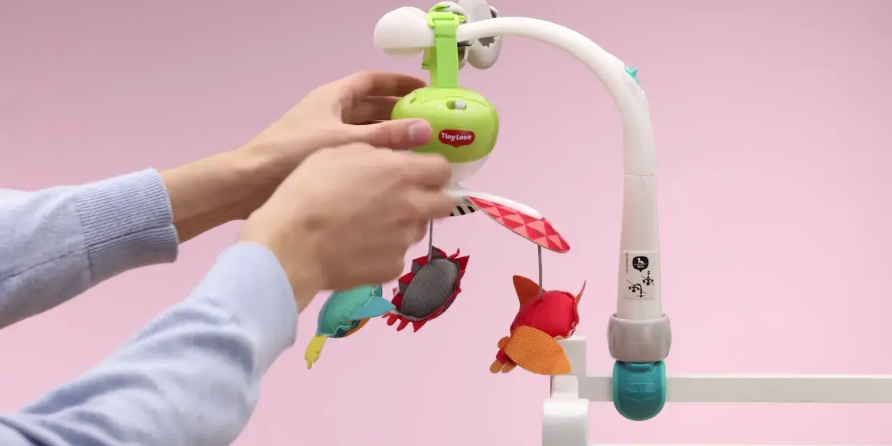 Ręce montują karuzelę podróżną Tiny Love z kolorowymi zabawkami.