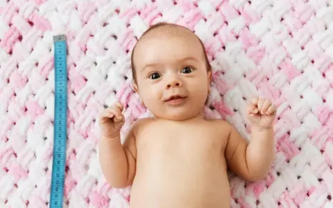 Niebieska miarka obok niemowlaka pokazuje, ile cm rośnie dziecko. Uroczy maluch leży na różowo-białym kocyku.