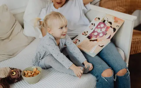 Mała dziewczynka z blond kucykami słucha bajki czytanej przez opiekunkę. Zastanawiasz się, ile zarabia opiekunka dla dziecka?