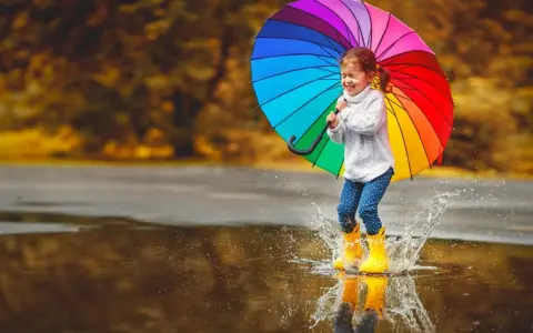 Radosna dziewczynka w żółtych kaloszach skacze po kałużach z tęczowym parasolem. Świetny pomysł, gdzie pojechać z dzieckiem, gdy pada deszcz!