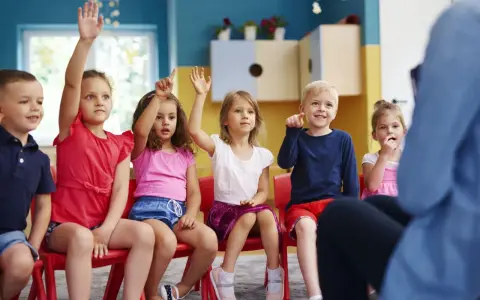 Dzieci na czerwonych krzesłach podnoszą ręce, odpowiadając na pytanie, ile lat ma się w zerówce.