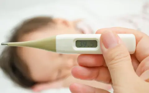 Cyfrowy termometr pokazuje 39.5°C. W tle widać śpiące dziecko. Zastanawiasz się, ile czasu mierzyć temperaturę pod pachą?