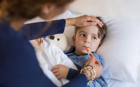 Chłopiec leży w łóżku z termometrem w ustach. Mama sprawdza mu temperaturę. Czy przy stanie podgorączkowym można iść na dwór?