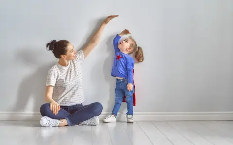 Mama mierzy wzrost córeczki w stroju superbohaterki. To właśnie wtedy dzieci rosną najszybciej, zaskakując nas każdego dnia.