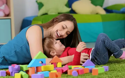 Uśmiechnięta opiekunka bawi się z dzieckiem wśród kolorowych klocków. Zastanawiasz się, ile kosztuje opieka nad dzieckiem za godzinę?