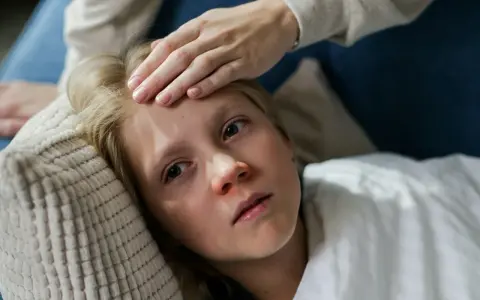 Dziecko z gorączką leży na poduszce, a ręka mamy sprawdza mu czoło. Czy dziecko z gorączką może wychodzić na dwór?