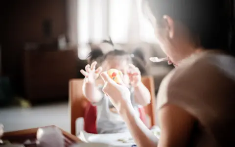 Niania karmi dziecko, które trzyma zabawkę. Zastanawiasz się, ile kosztuje godzina niani do dziecka?