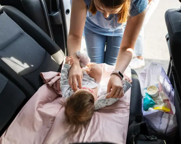 Mama uczy się, jak przebierać noworodka w samochodzie. Na różowej macie leży dziecko, a obok torba z akcesoriami.