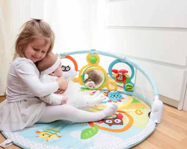 Starsza siostra tuli niemowlę na kolorowych matach interaktywnych dla dzieci, pełnych zabawek i lusterka.