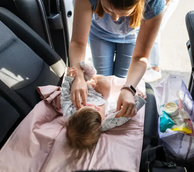 Mama uczy się, jak przebierać noworodka w samochodzie. Na różowej macie leży dziecko, a obok torba z akcesoriami.