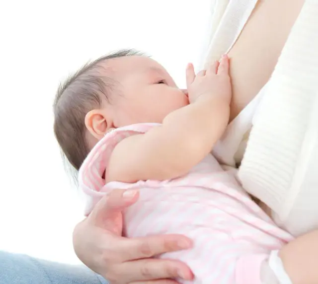 Malutkie dziecko karmione piersią. Mama delikatnie przytrzymuje główkę, a dziecko chwyta pierś. Nie widać kapturków na brodawki.