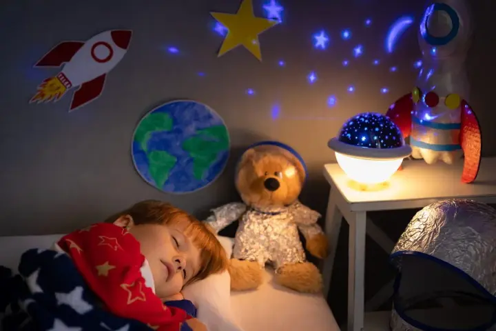 Śpiące dziecko pod kocem w gwiazdki, obok pluszowy miś w stroju astronauty. Na ścianie rakieta i Ziemia, a na suficie projektor dla dzieci na sufit wyświetla gwiazdy.
