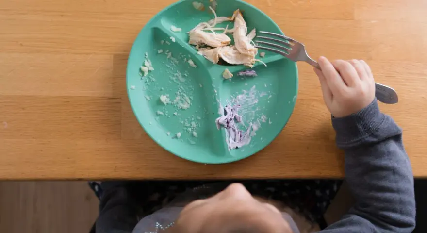 Dziecko je kurczaka z talerza babyono z przyssawką. Resztki jedzenia na talerzu i widoczna rączka z widelcem.