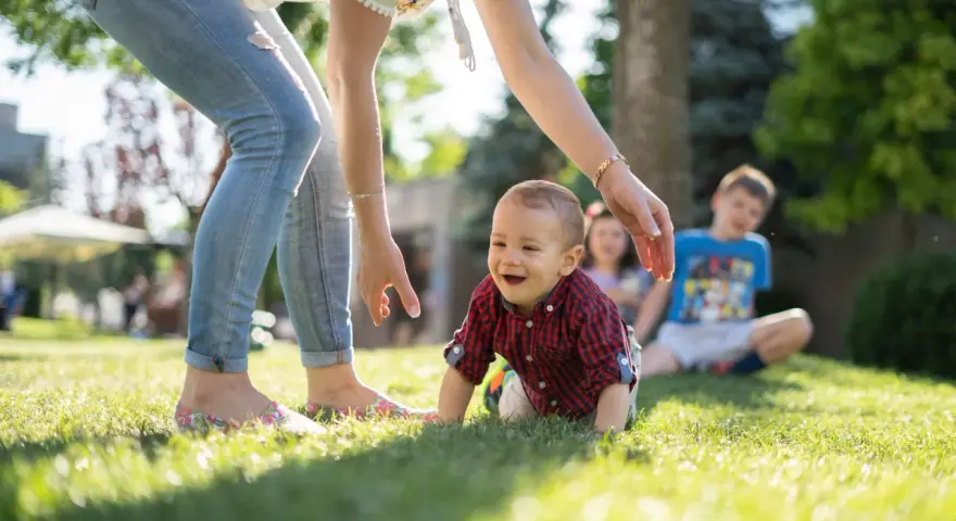 Uśmiechnięte dziecko raczkuje na trawie, podczas gdy opiekunka zastanawia się, ile kosztuje niania na pełen etat.