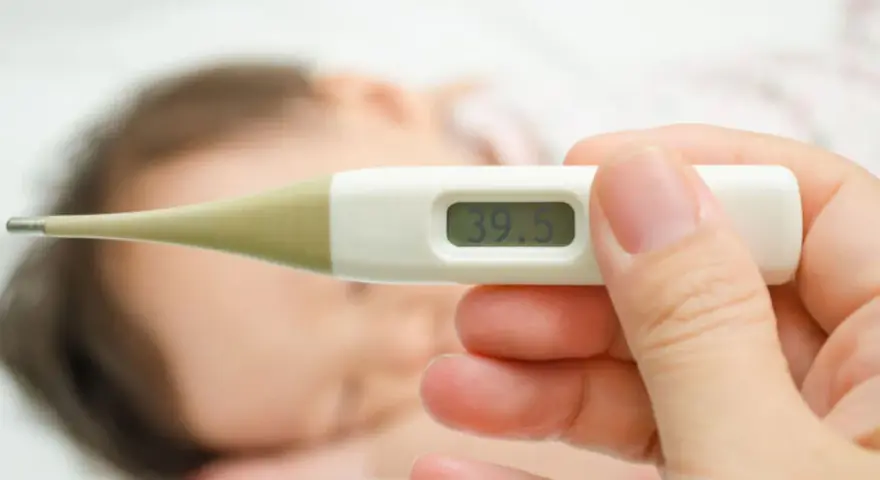 Cyfrowy termometr pokazuje 39.5°C. W tle widać śpiące dziecko. Zastanawiasz się, ile czasu mierzyć temperaturę pod pachą?