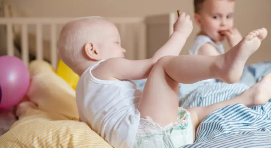 Dzieci bawią się na łóżku. Maluch w pieluszce sięga po zabawkę, a starszy chłopiec je. Ciekawe, ile pieluch miesięcznie zużywają takie energiczne maluchy!