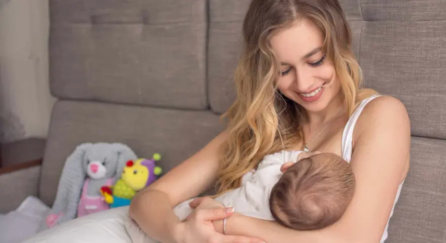 Uśmiechnięta mama karmi piersią dziecko. W tle pluszowe zabawki. Nakładki na pierś do karmienia mogą pomóc w tym intymnym momencie.