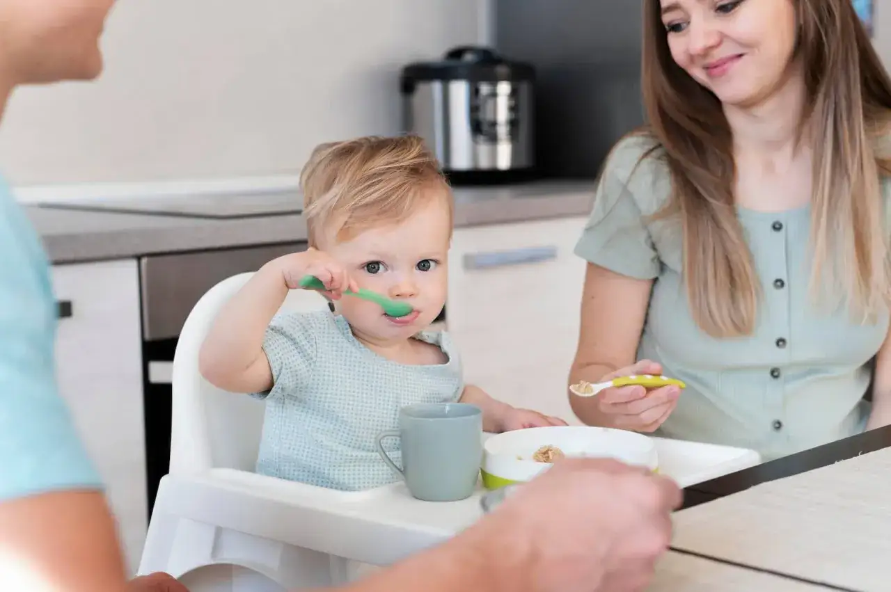 Małe dziecko uczy się jeść samodzielnie, używając zielonej łyżeczki do rozszerzania diety. Mama z uśmiechem podaje mu kolejne porcje.