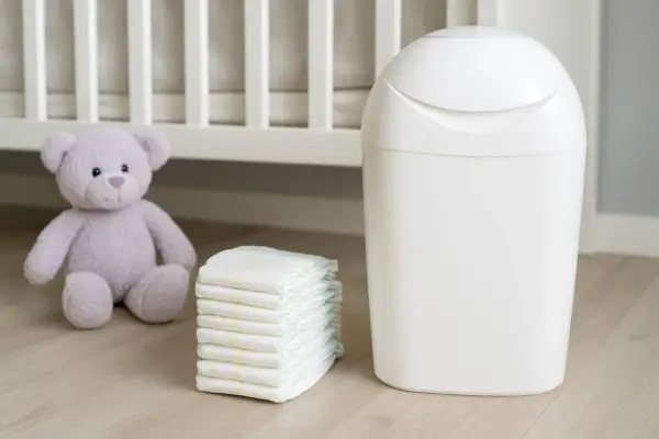 Biały kosz na pampersy Angelcare obok stosu pieluch i pluszowego misia, gotowy do utrzymania czystości w pokoju dziecka.