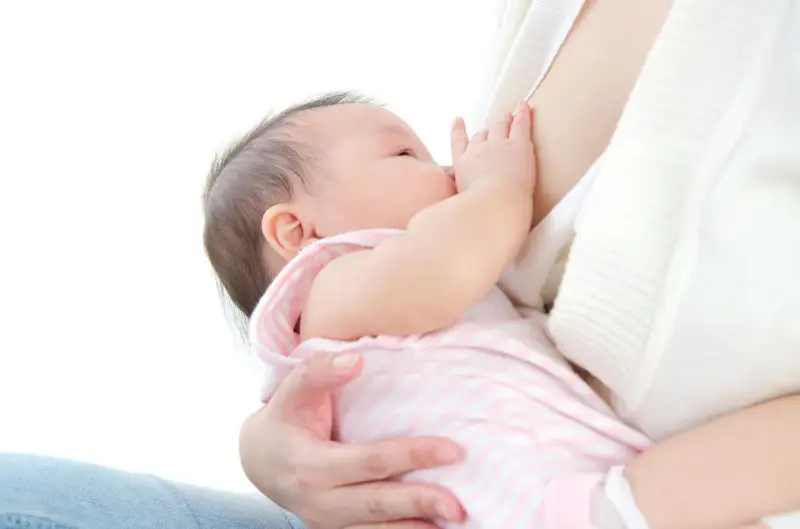 Malutkie dziecko karmione piersią. Mama delikatnie przytrzymuje główkę, a dziecko chwyta pierś. Nie widać kapturków na brodawki.