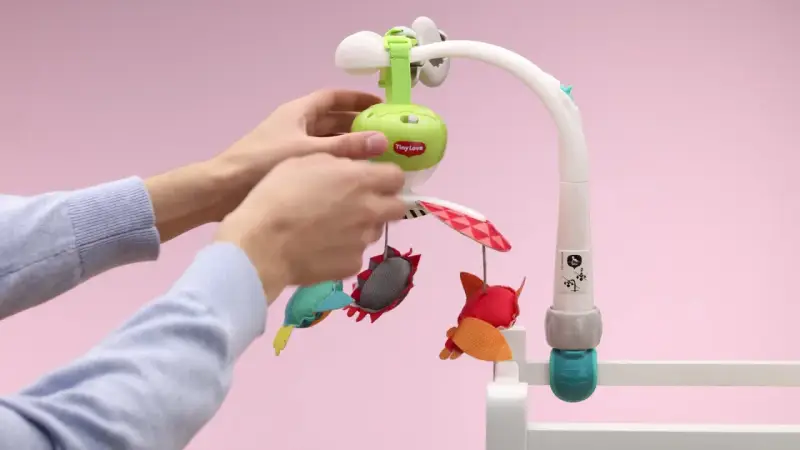 Ręce montują karuzelę podróżną Tiny Love z kolorowymi zabawkami.